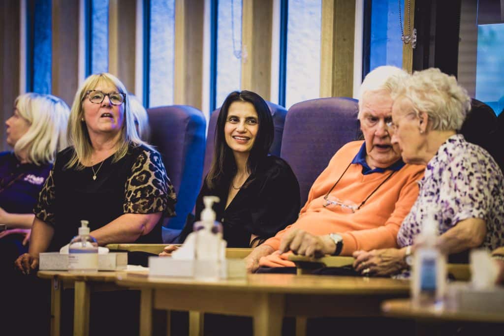 Group of older adults seated along a wooden table, chatting; left woman in leopard print sleeves, center smiling woman in black, two seniors on the right in conversation.