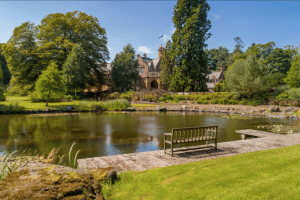 Stone manor surrounded by trees across a calm pond; a wooden dock and bench sit in the foreground.