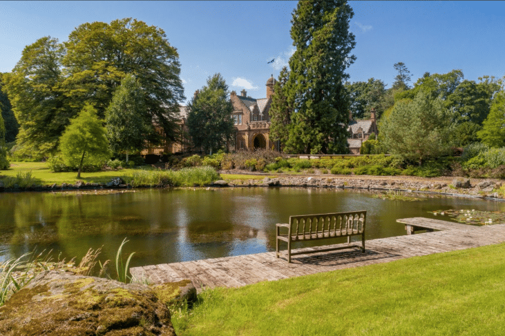 Stone manor surrounded by trees across a calm pond; a wooden dock and bench sit in the foreground.