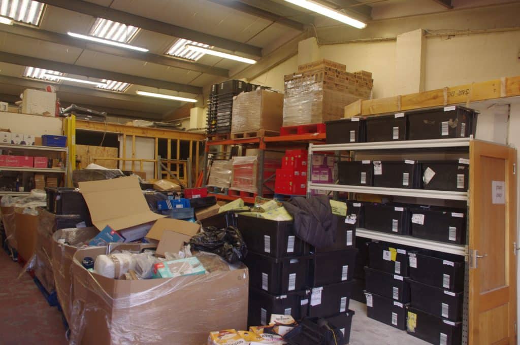 Inside a cluttered warehouse with shelves, pallets, and stacked boxes and crates along the walls.