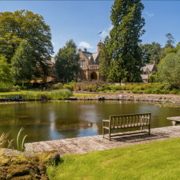 Stone manor surrounded by trees across a calm pond; a wooden dock and bench sit in the foreground.